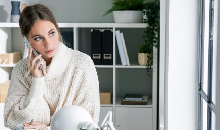 Moteris kalba mobiliuoju telefonu žiūrėdama pro langą moderniame namų biure su lentynomis fone
