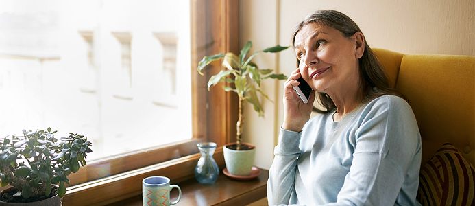 Mobiliojo ryšio pokalbių planai 4 Vyresnio amžiaus moteris sėdi prie lango jaukiame kambaryje, kalba telefonu ir žvelgia į šoną; ant palangės stovi kambariniai augalai, puodelis ir vaza, pro langą sklinda šilta dienos šviesa.