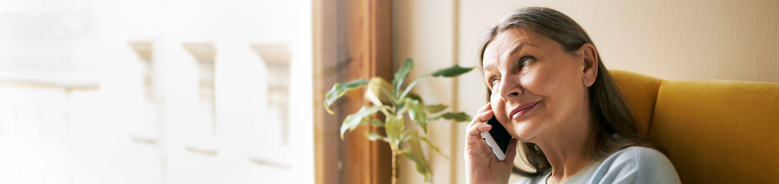 Vyresnio amžiaus moteris sėdi prie lango jaukiame kambaryje, kalba telefonu ir žvelgia į šoną; ant palangės stovi kambariniai augalai, puodelis ir vaza, pro langą sklinda šilta dienos šviesa.