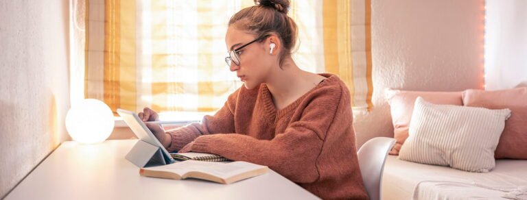 Jauna mergina sėdi prie stalo ir mokosi: naudoja planšetinį kompiuterį su rašikliu, šalia guli atversta knyga; ji dėvi akinius ir belaides ausines, kambarį apšviečia šilta šviesa.